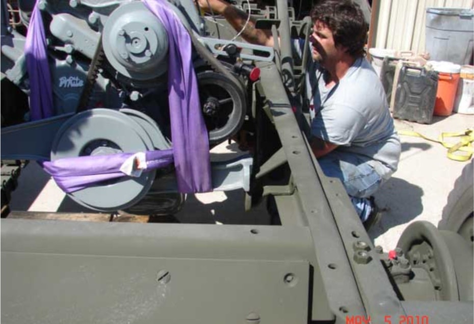 Man in gray shirt working on engine being lowered into halftrack with purple straps